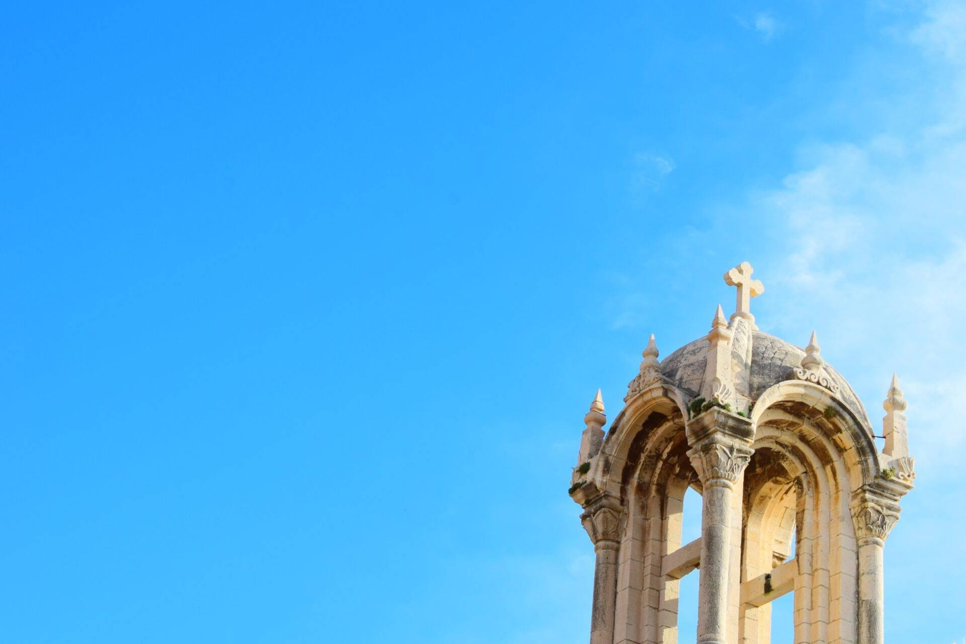 Capilla cristiana en ruinas con columnas ornamentadas y cruz dorada en lo alto, cielo azul despejado