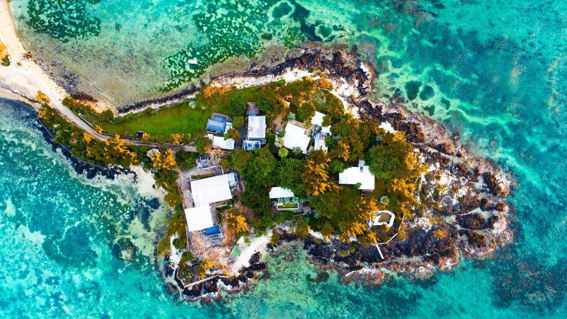 Vista aérea de una pequeña isla rocosa con casas blancas, vegetación verde y aguas turquesas cristalinas