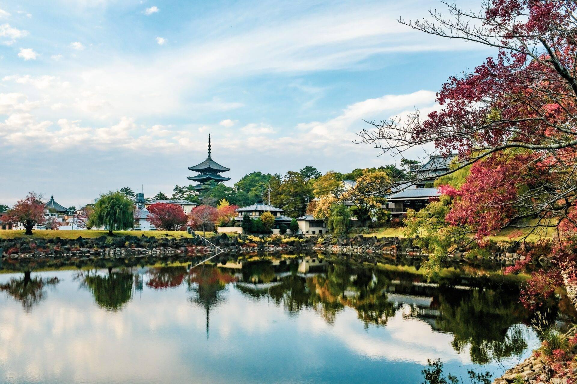 Japanische Herbstlandschaft mit einem traditionellen Tempel, der sich in einem ruhigen Fluss spiegelt, umgeben von Bäumen mit roten und goldenen Blättern