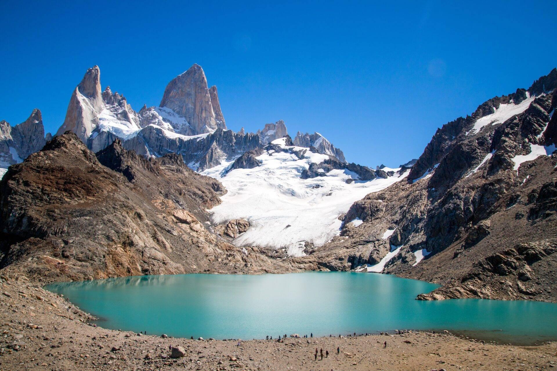 Lago glaciale turchese incorniciato da cime rocciose innevate e un ghiacciaio bianco sotto un cielo limpido