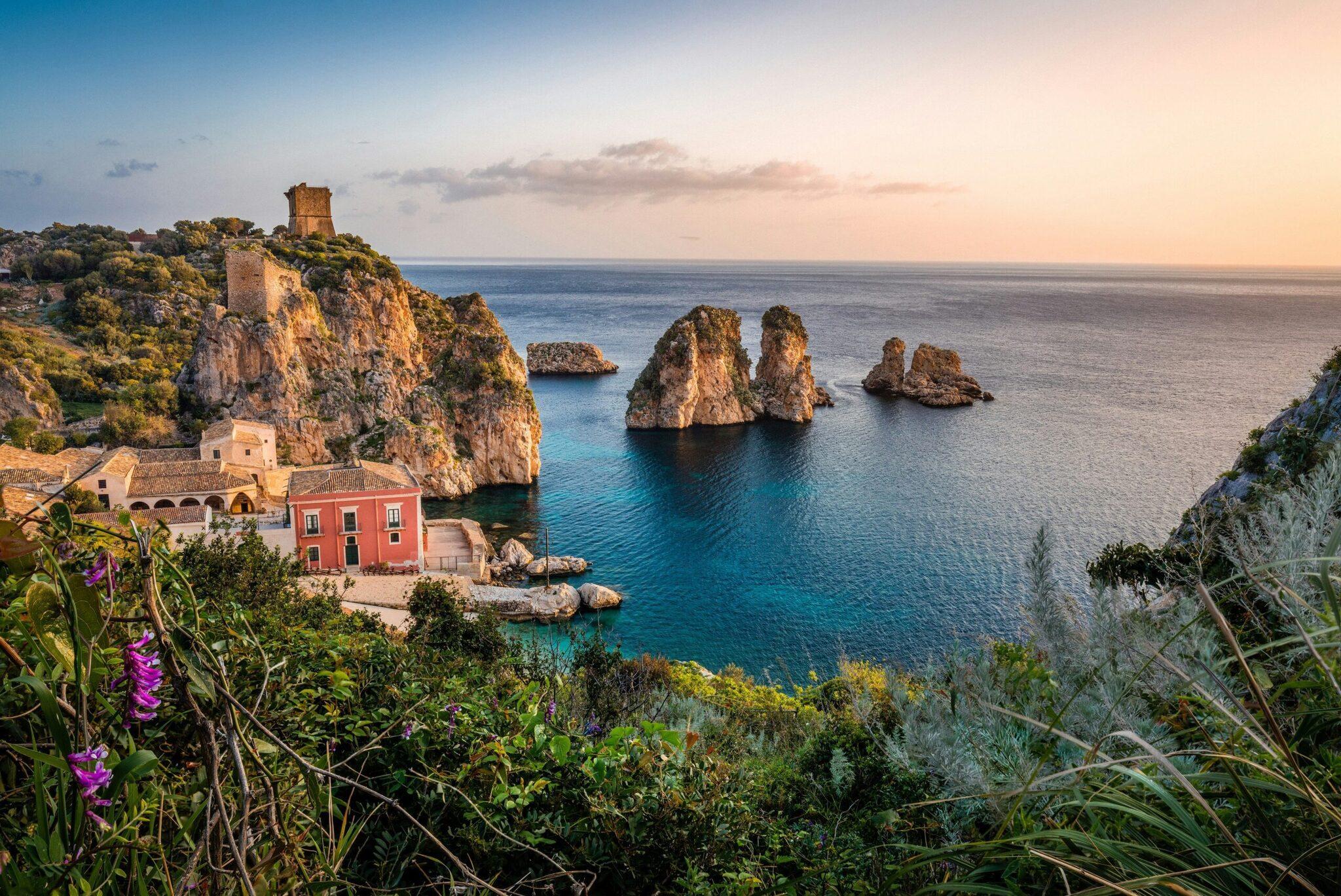Vista costiera mediterranea con case colorate, falesie rocciose e formazioni geologiche in mare al tramonto