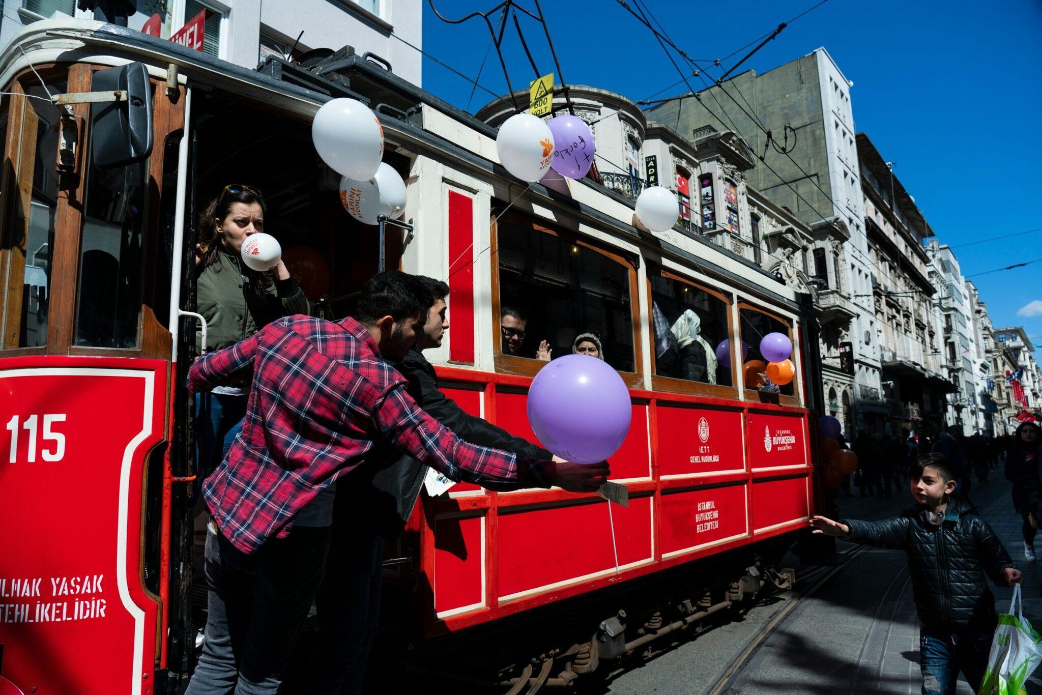 Un tram rosso storico decorato con palloncini bianchi e viola percorre una strada animata di una città