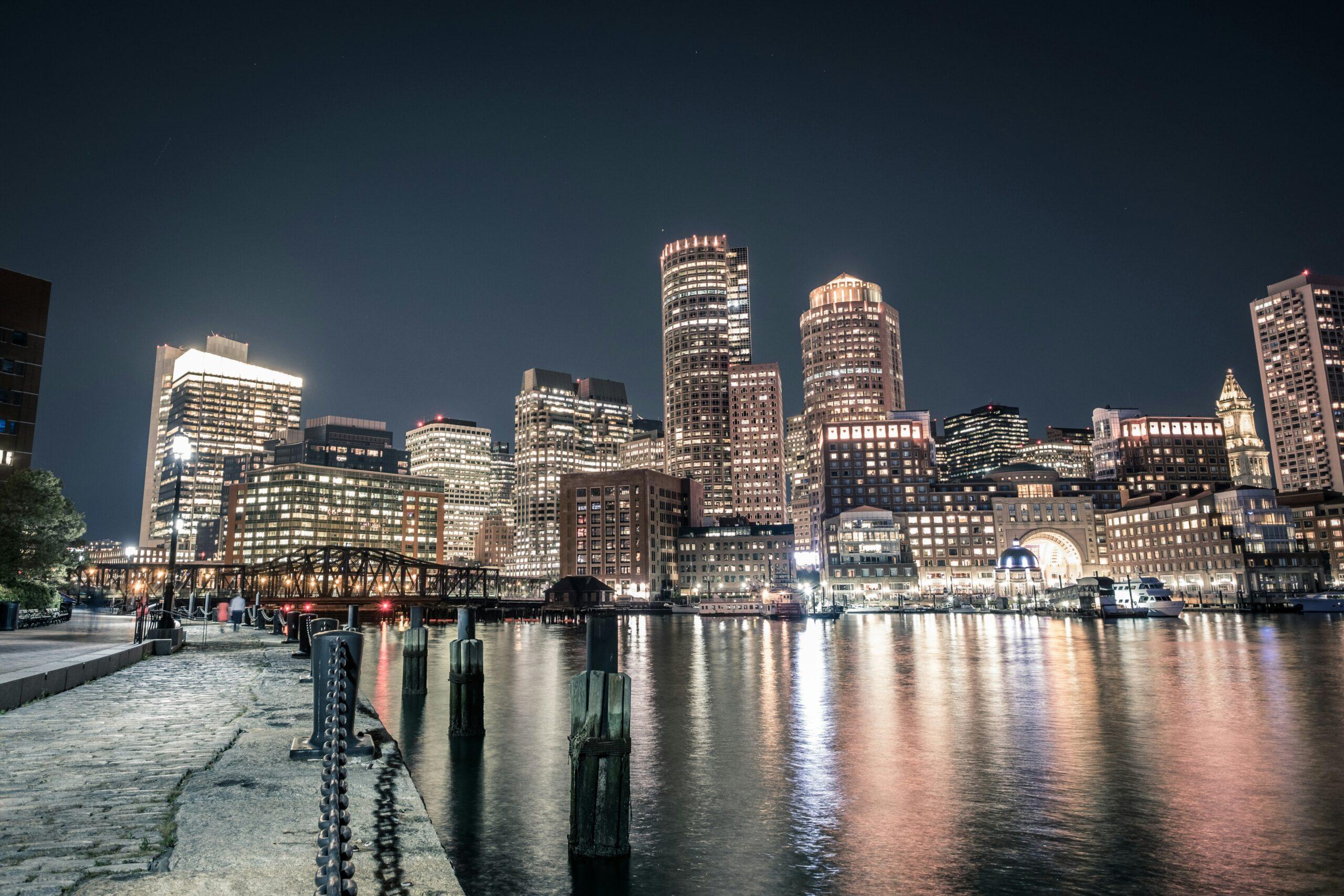 The illuminated skyline of a large coastal city at night, with skyscrapers and a metal bridge bordering a bay