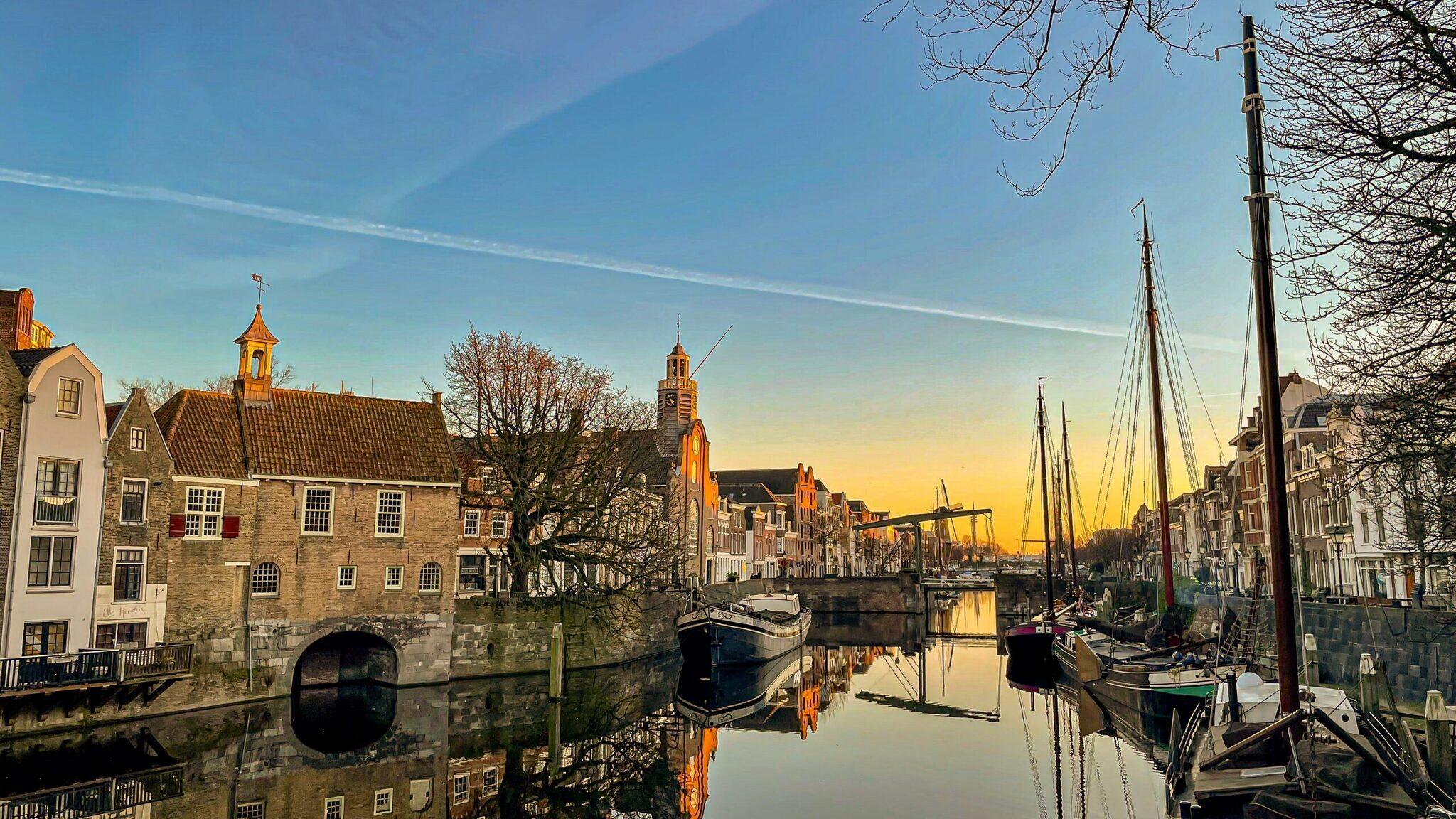 Historischer Kanal mit traditionellen Häusern, vertäuten Segelschiffen und Kirchen bei Sonnenuntergang in einer niederländischen Hafenstadt