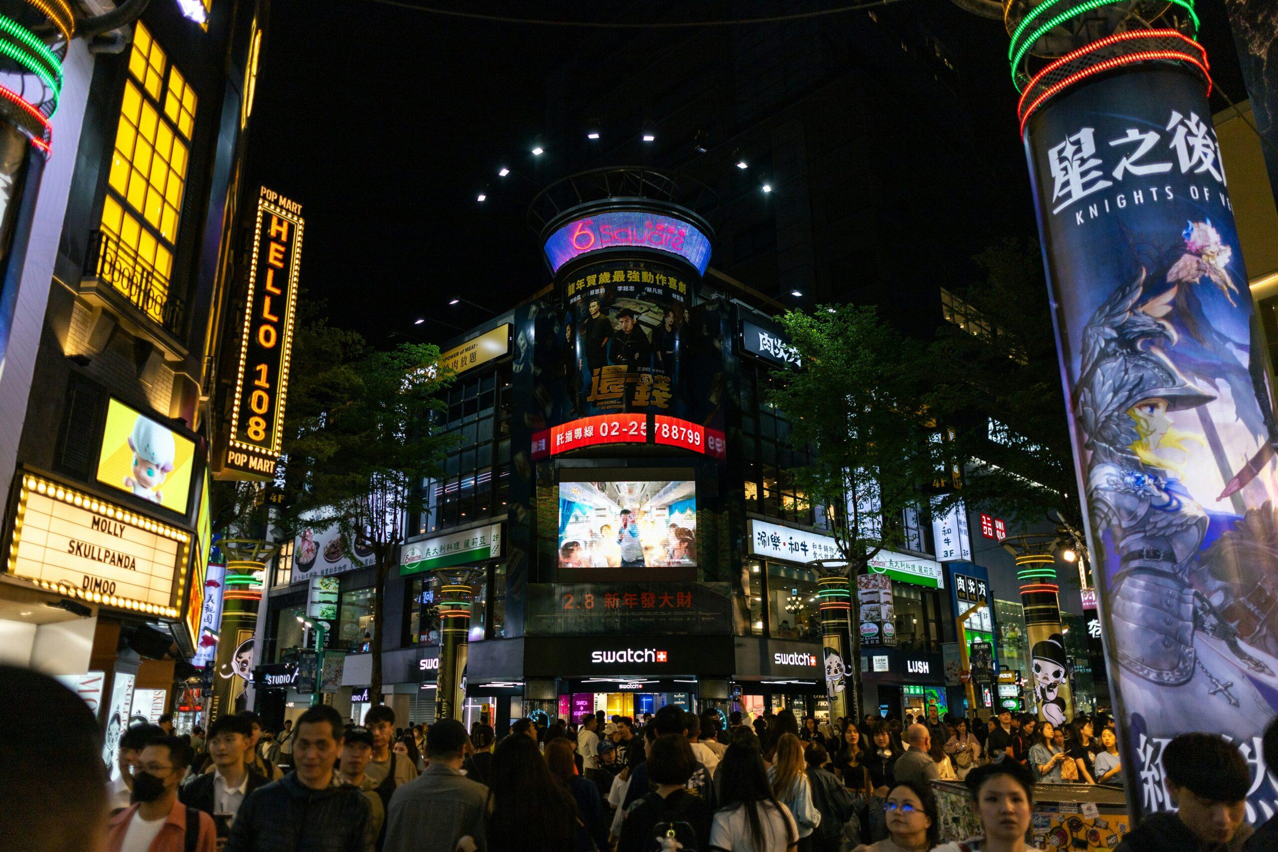 Calle comercial animada de noche con letreros luminosos, pantallas gigantes y multitud de peatones en un barrio urbano asiático