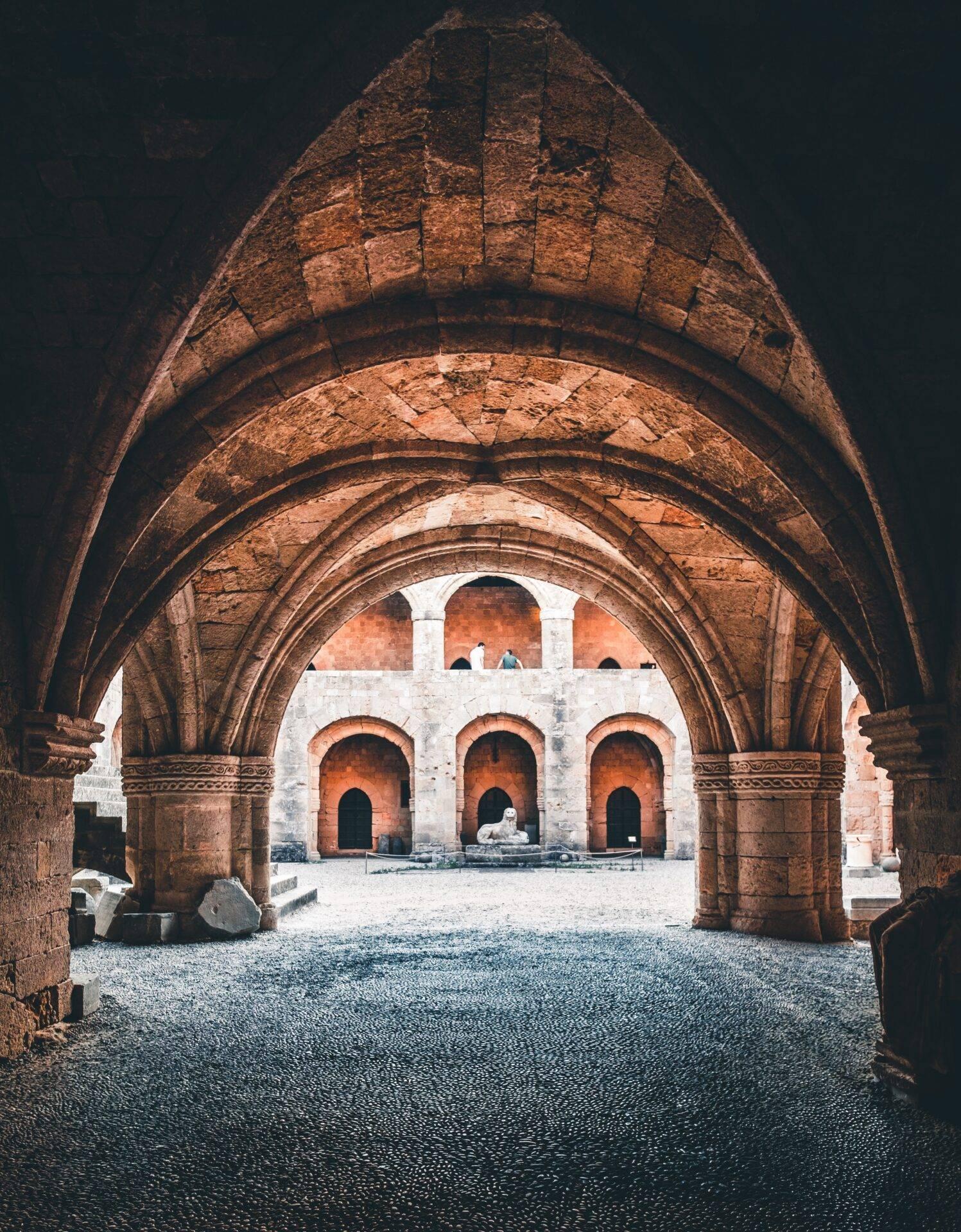 Blick durch einen gewölbten roten Steinbogen auf einen Innenhof mit drei weißen und ockerfarbenen Arkaden