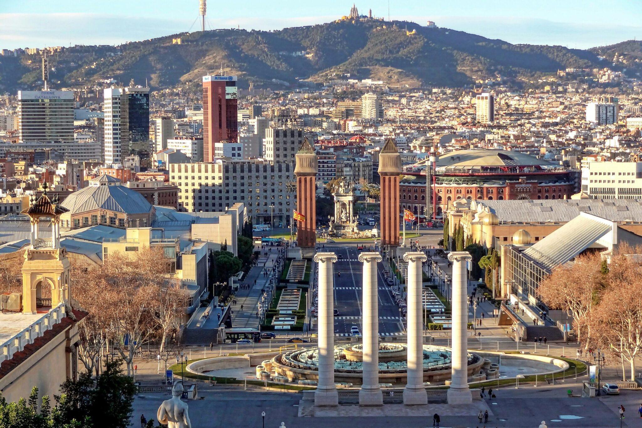 Vista aérea de una plaza con cuatro columnas monumentales, edificios históricos y una colina arbolada al fondo