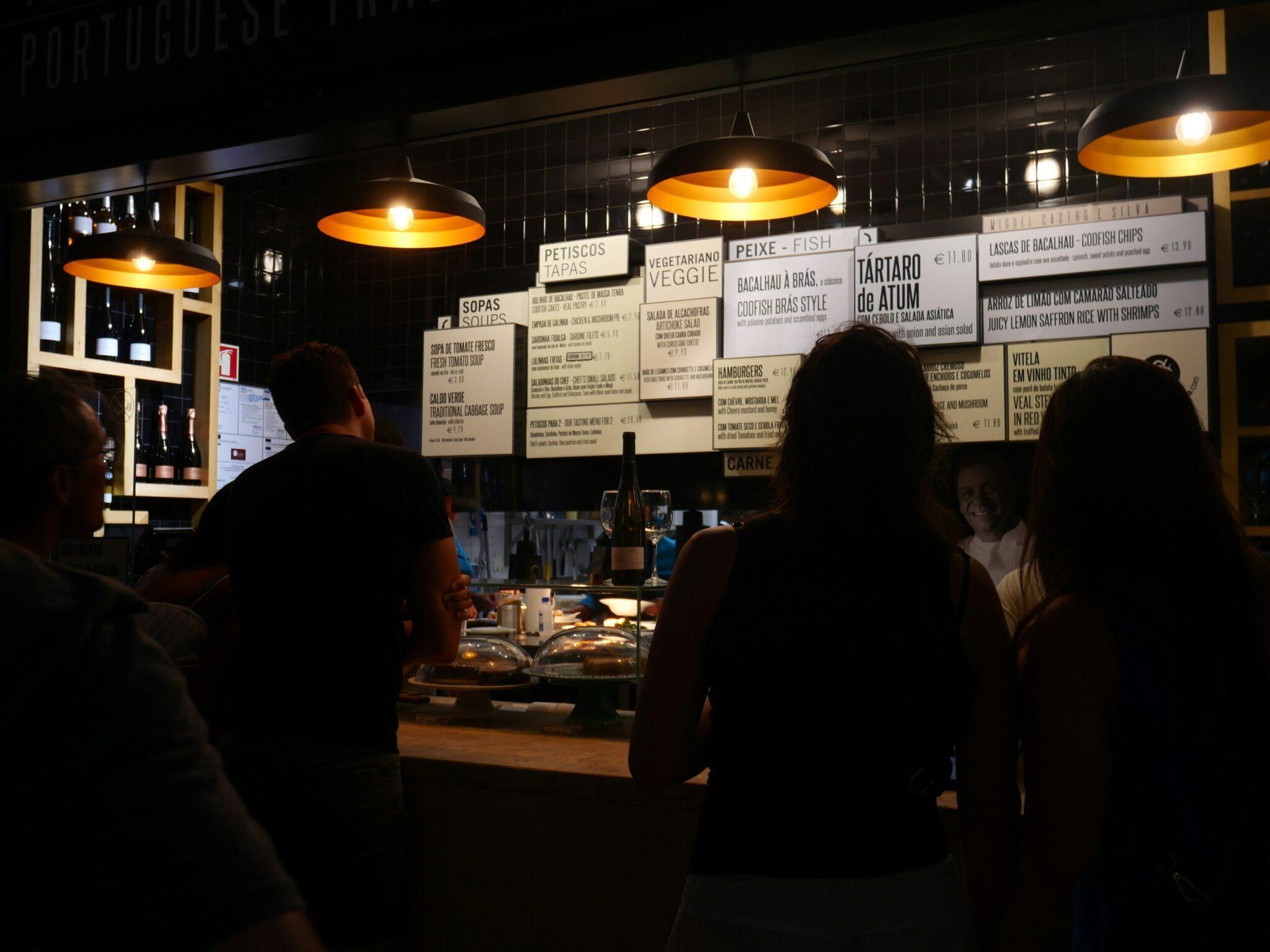 Eine kleine portugiesische Taverne bei Nacht mit ausgehängter Speisekarte, Gästen an der Theke und warmem Lampenlicht