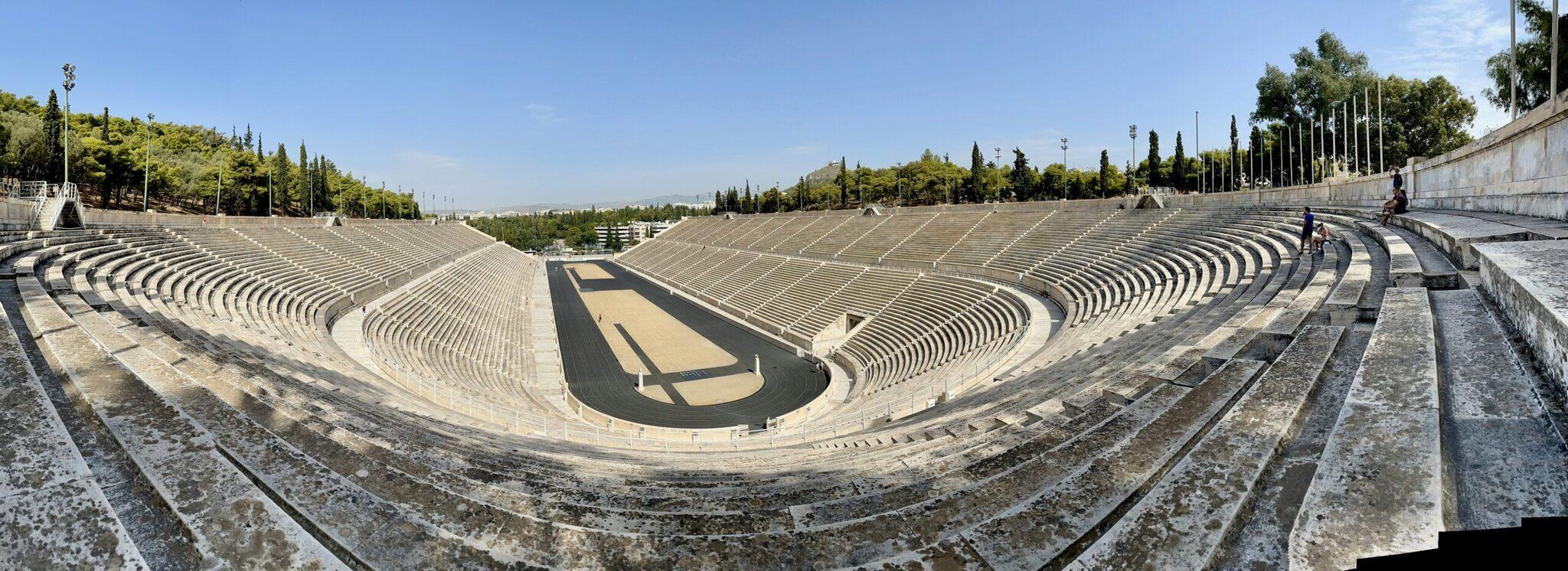 Ein antikes Amphitheater-Stadion aus weißem Marmor mit konzentrischen Rängen unter klarem Himmel, umgeben von Zypressen
