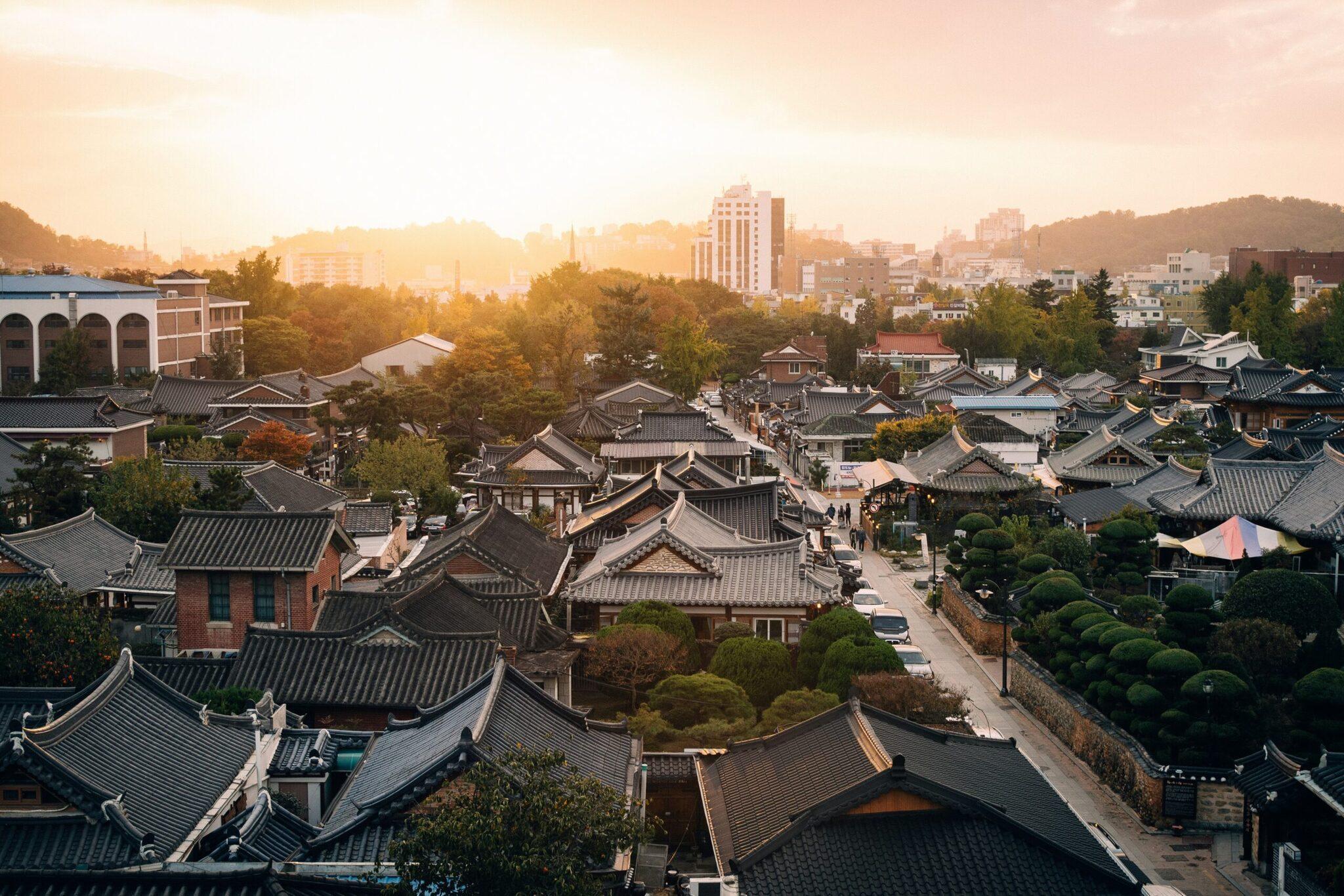 Vogelperspektive auf ein traditionelles koreanisches Dorf mit geschwungenen Ziegeldächern, umgeben von modernen Wolkenkratzern bei Sonnenuntergang