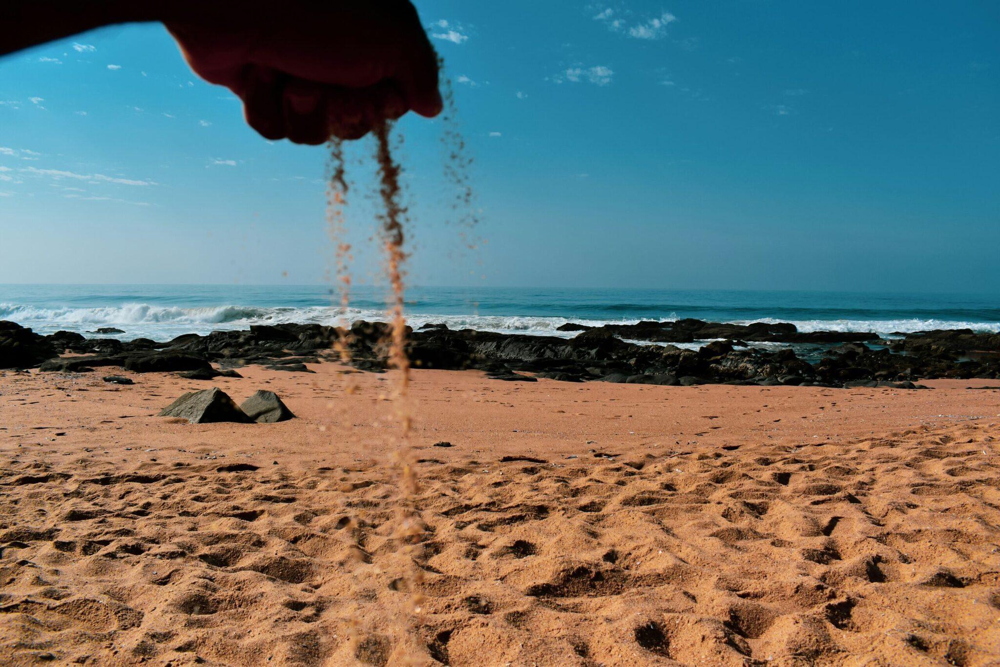 Una mano deja caer arena fina en una playa rocosa con vistas al océano y las olas