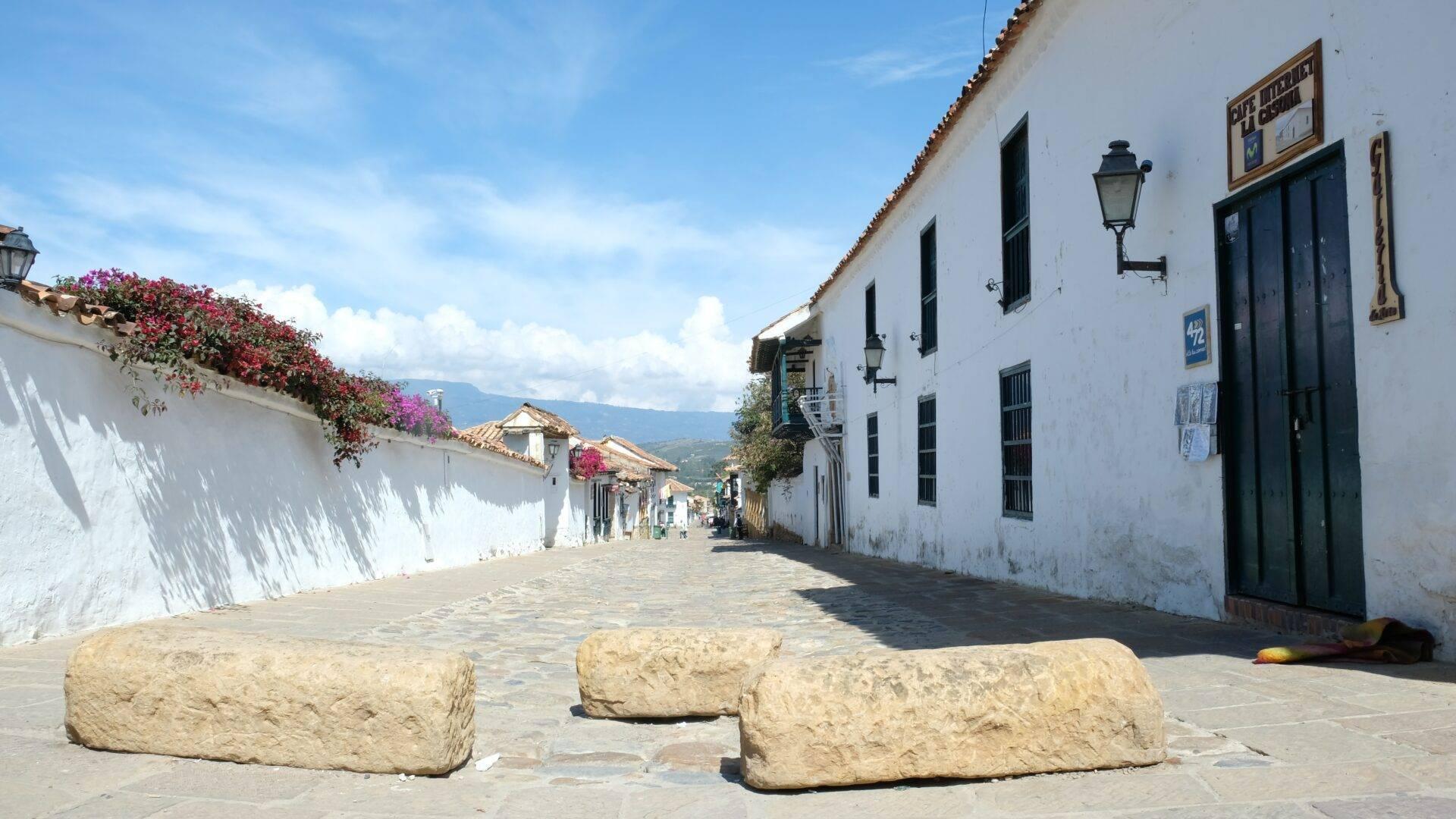 Calle empedrada pintoresca flanqueada por casas coloniales blancas con flores rosas y montañas al fondo