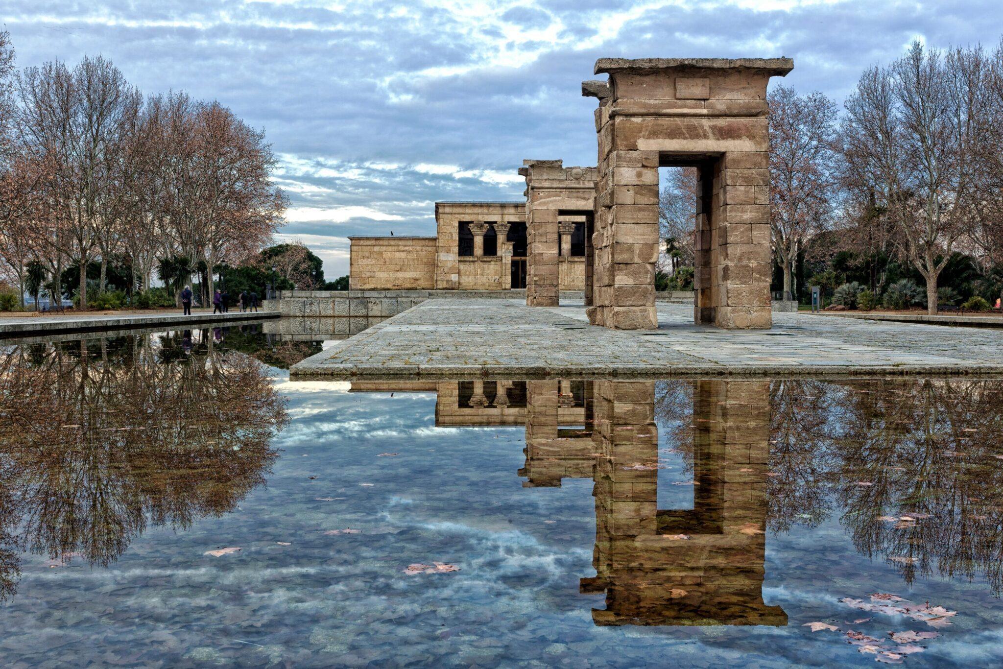Le rovine di un tempio antico si riflettono in uno specchio d’acqua tranquillo, circondato da alberi spogli e un cielo nuvoloso