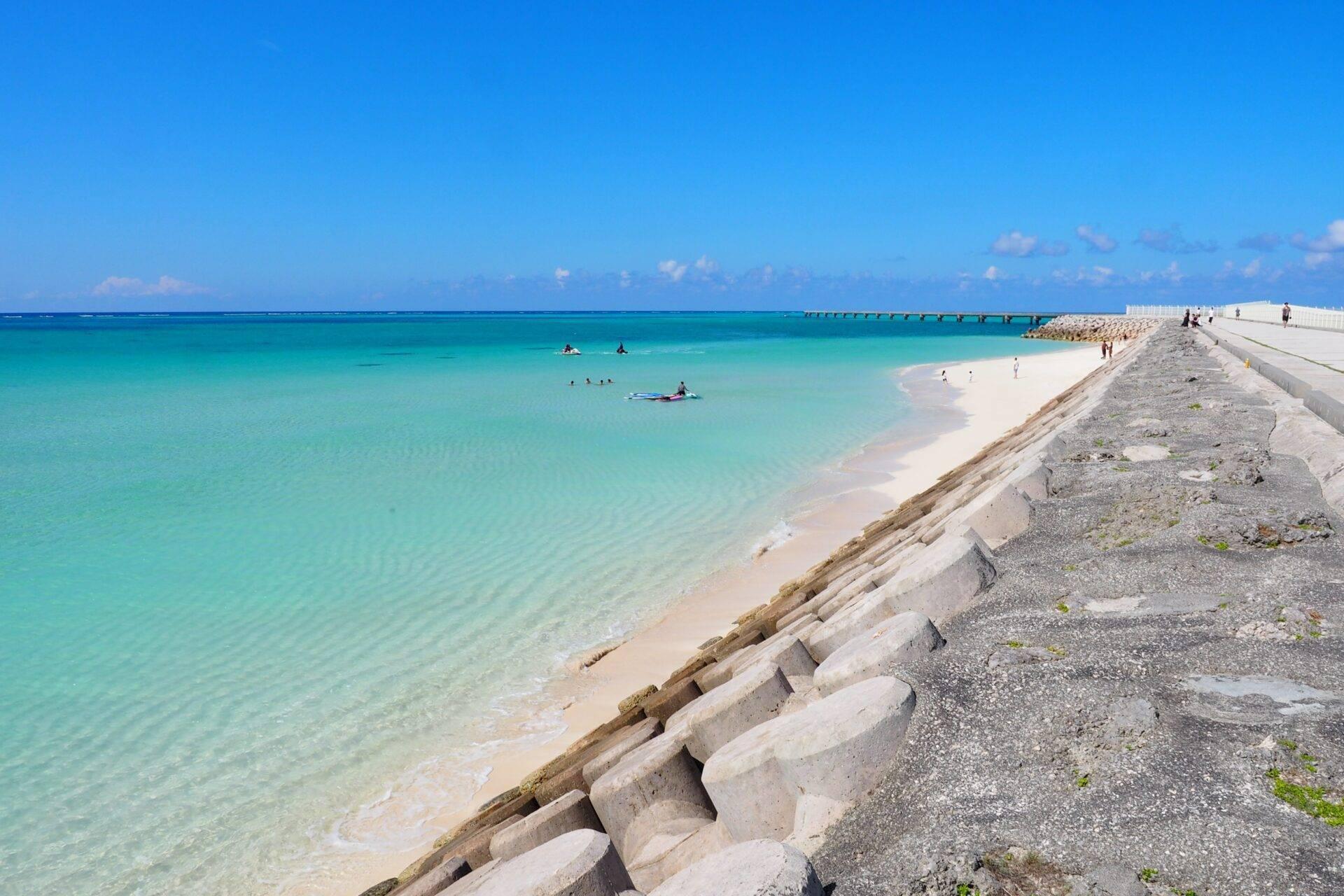 Playa de arena blanca con aguas turquesas cristalinas, dique de hormigón y bañistas